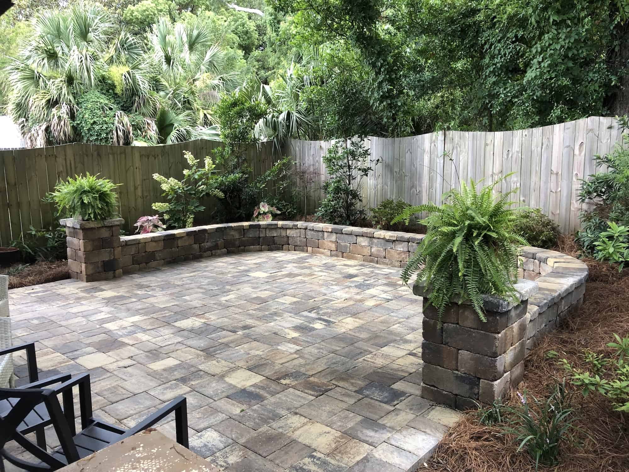 Backyard stone patio with brick/stone wall surrounded by various potted/planted plants in straw
