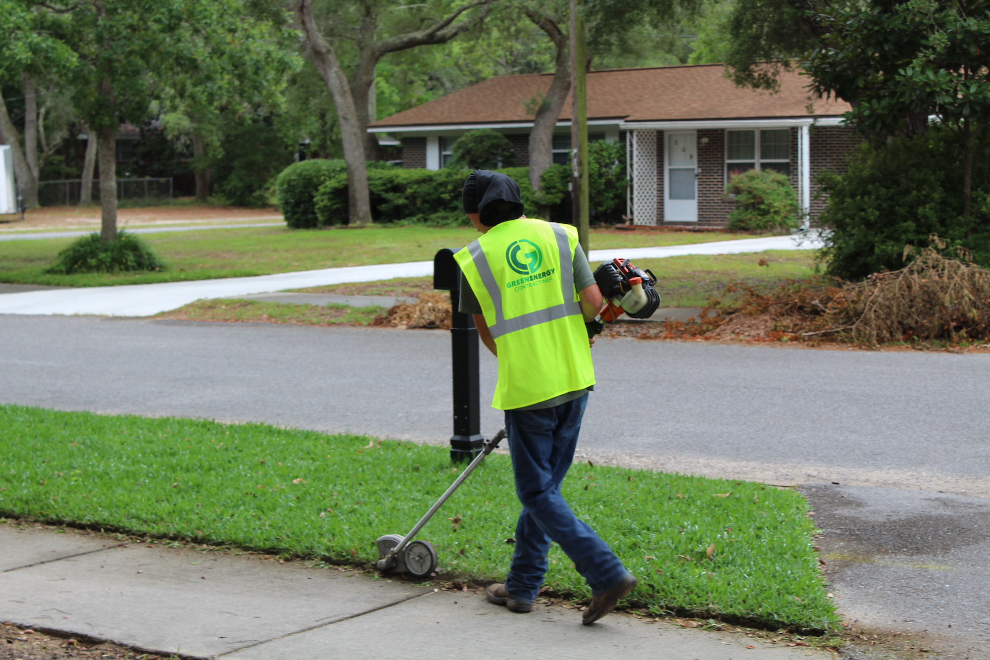 Green Energy Landscaping employee edge trimming lawn against sidewalk, wearing bright green safety vest with GEL logo