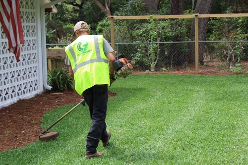 Green Energy Landscaping employee string trimming green grass up against red mulch on the side of a white breeze block house