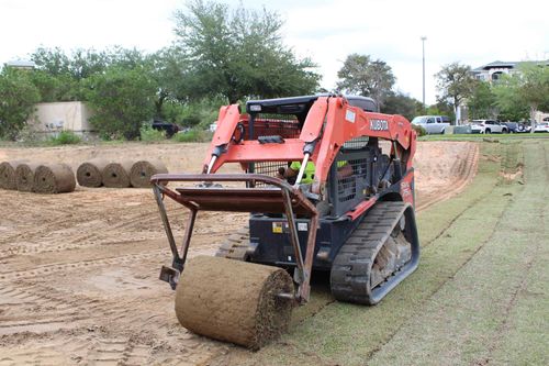 Big roll track installer rolling out sod over dirt. Next to it are lines of rolled out sod and on the other side, rolls of sod yet to be rolled out
